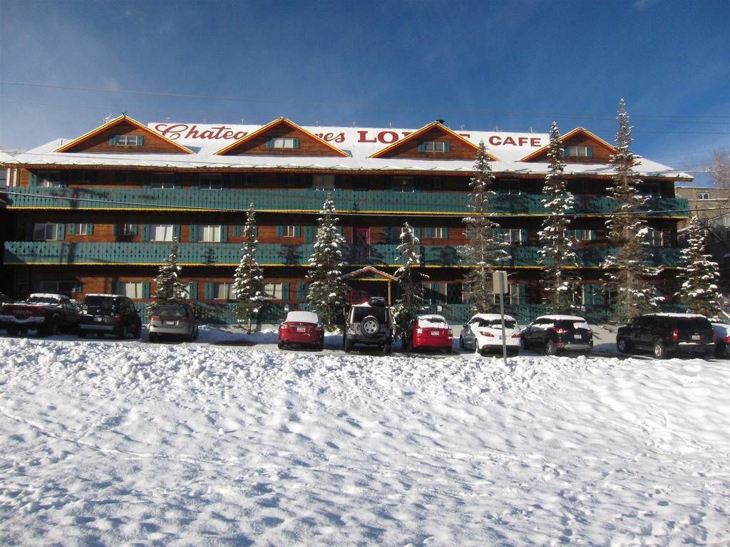 A hotel with a snow-covered roof and multiple balconies. There are several cars parked in front, and snow-covered trees are visible.