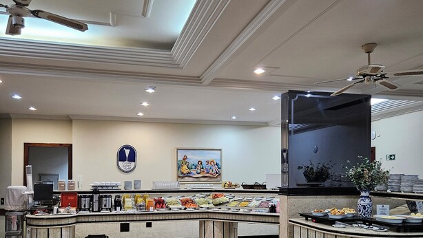 Un comedor con mesas redondas cubiertas con manteles rosas, una barra de buffet con diversos alimentos y ventiladores de techo.