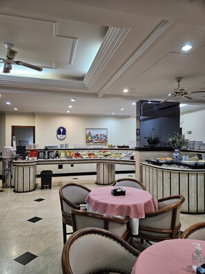 A dining area with round tables covered with pink tablecloths, a buffet counter with various food items, and ceiling fans. - Cristal Palace Hotel (Lins)