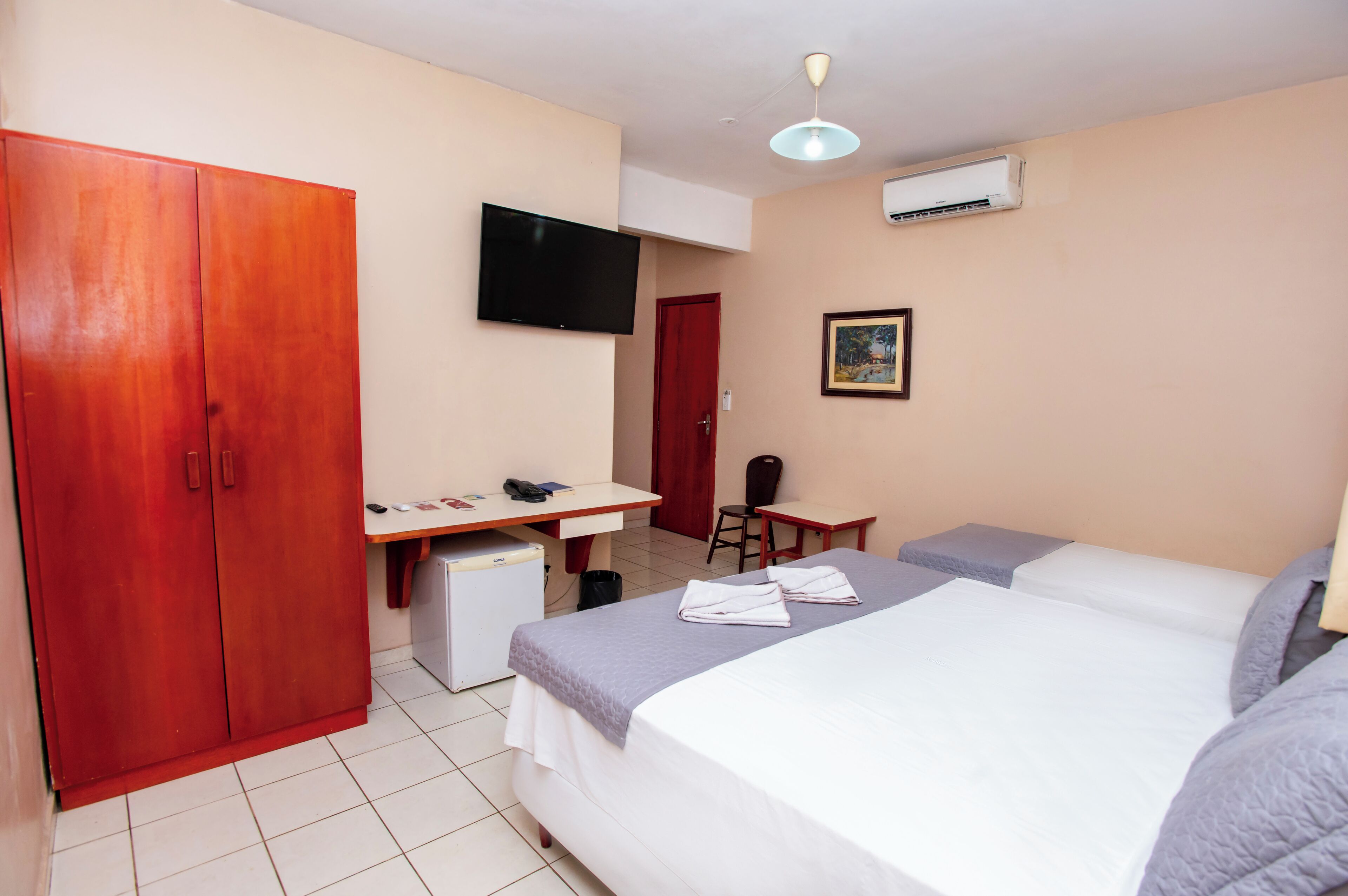 A hotel room with a bed, a desk, a wardrobe, a television, and an air conditioning unit.