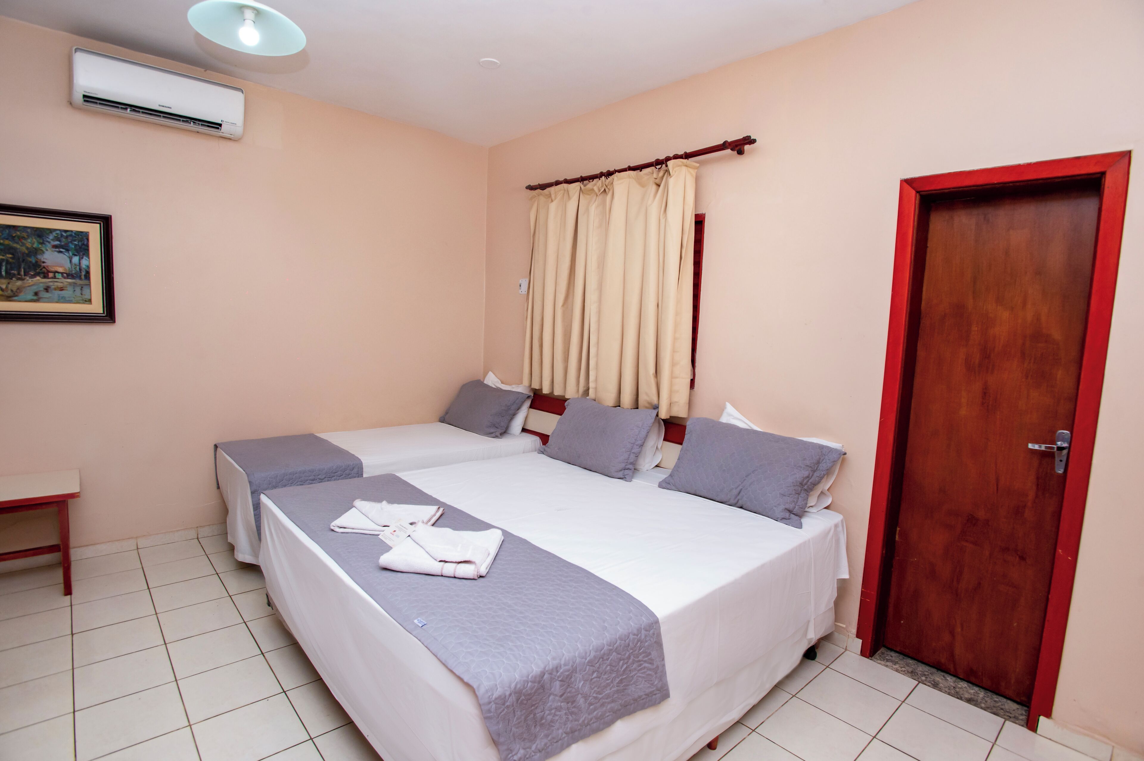 Quarto de hotel com duas camas, porta de madeira, ar condicionado branco e um quadro na parede.