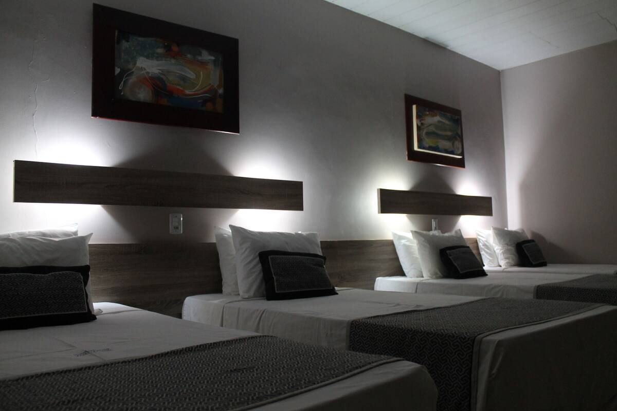 A hotel room with three beds, each with a headboard and pillows. There are wall-mounted light fixtures above each bed.