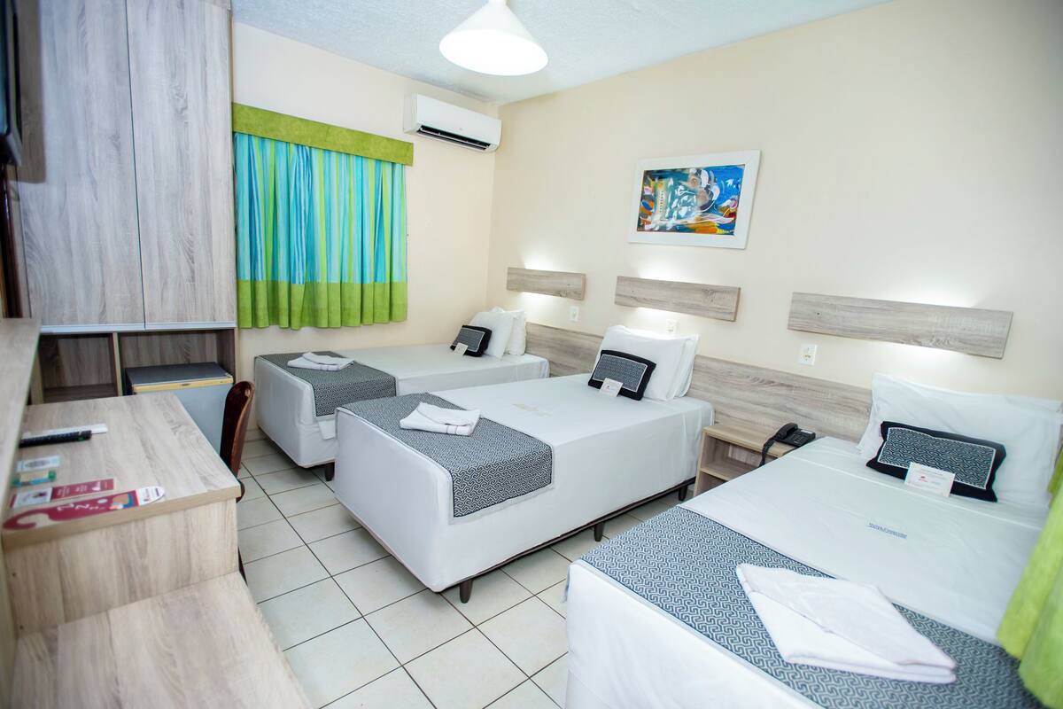 A hotel room with two beds, a desk, and a chair. There is a window with curtains, a painting on the wall, and an air conditioning unit.