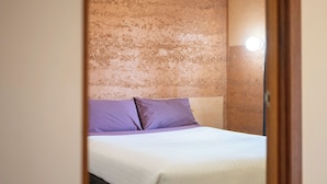 A bed with white bedding and two purple pillows in a room with a textured beige wall. - Mud Hut Motel (Coober Pedy)