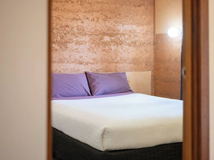 Una cama con ropa de cama blanca y dos almohadas moradas en una habitación con pared beige con textura.