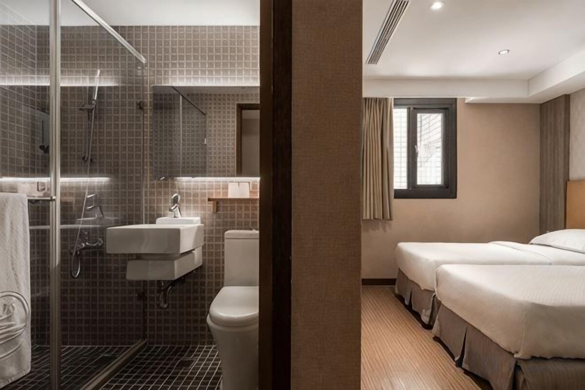 A hotel bathroom with a shower, sink, and toilet. There is a bed with a window in the adjacent room.