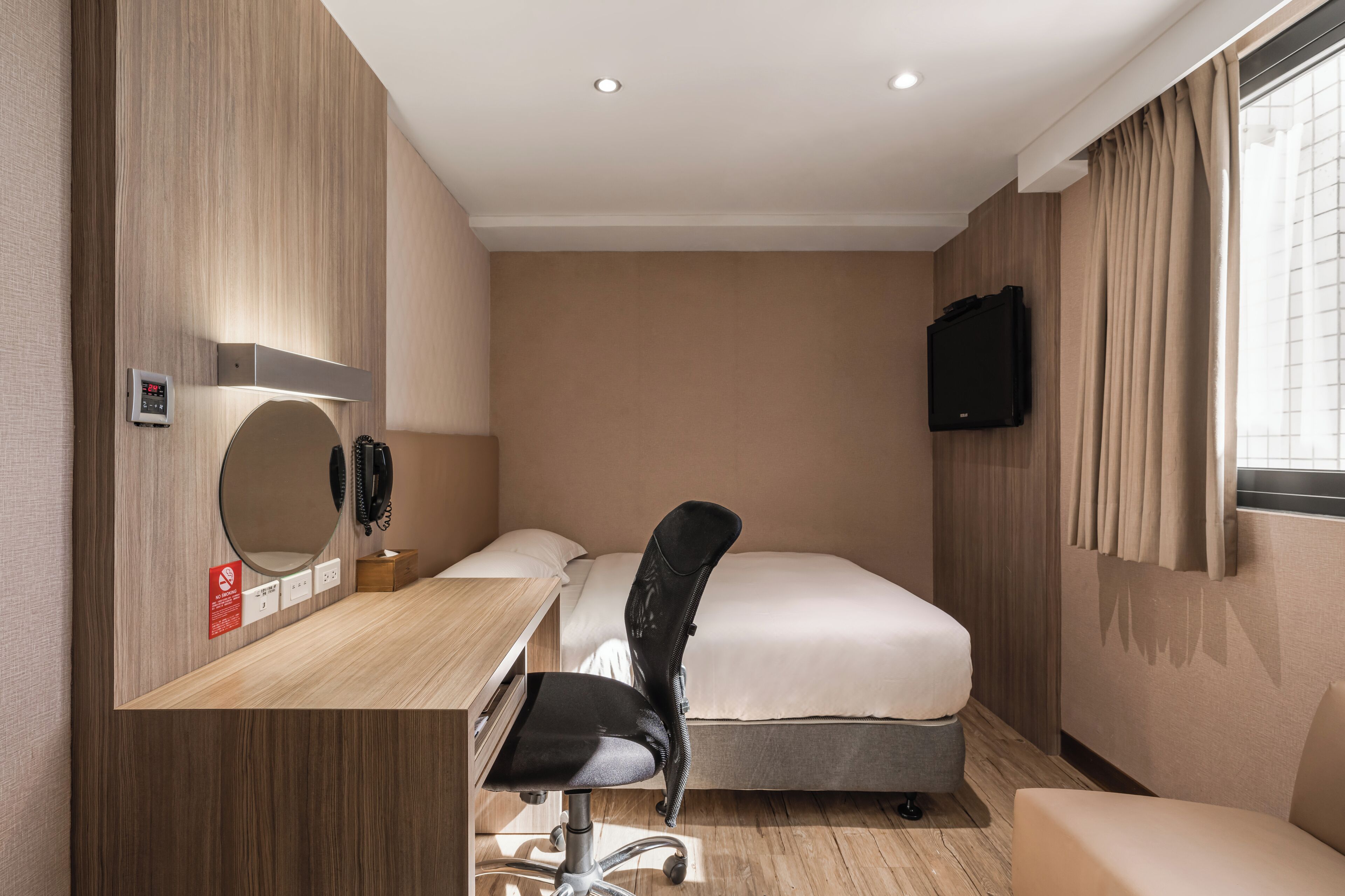 A hotel room with a bed, a desk with a chair, a TV mounted on the wall, and a window with curtains.