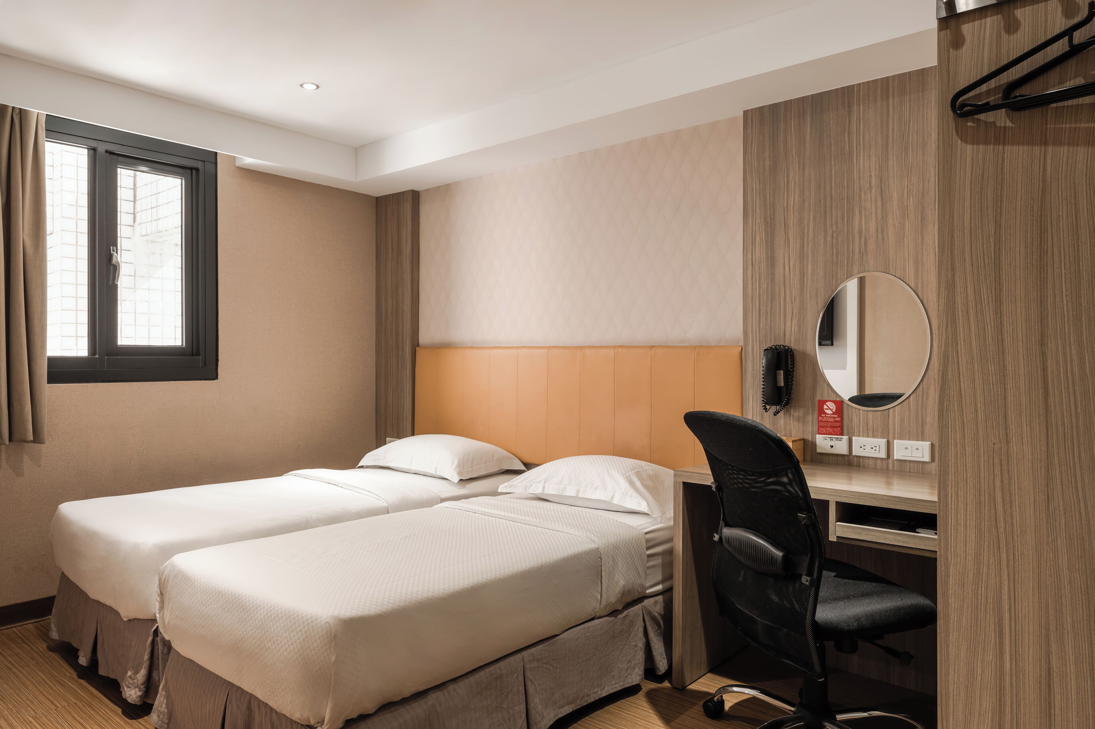 A hotel room with two single beds, a desk with a chair, a mirror, and a window with curtains.