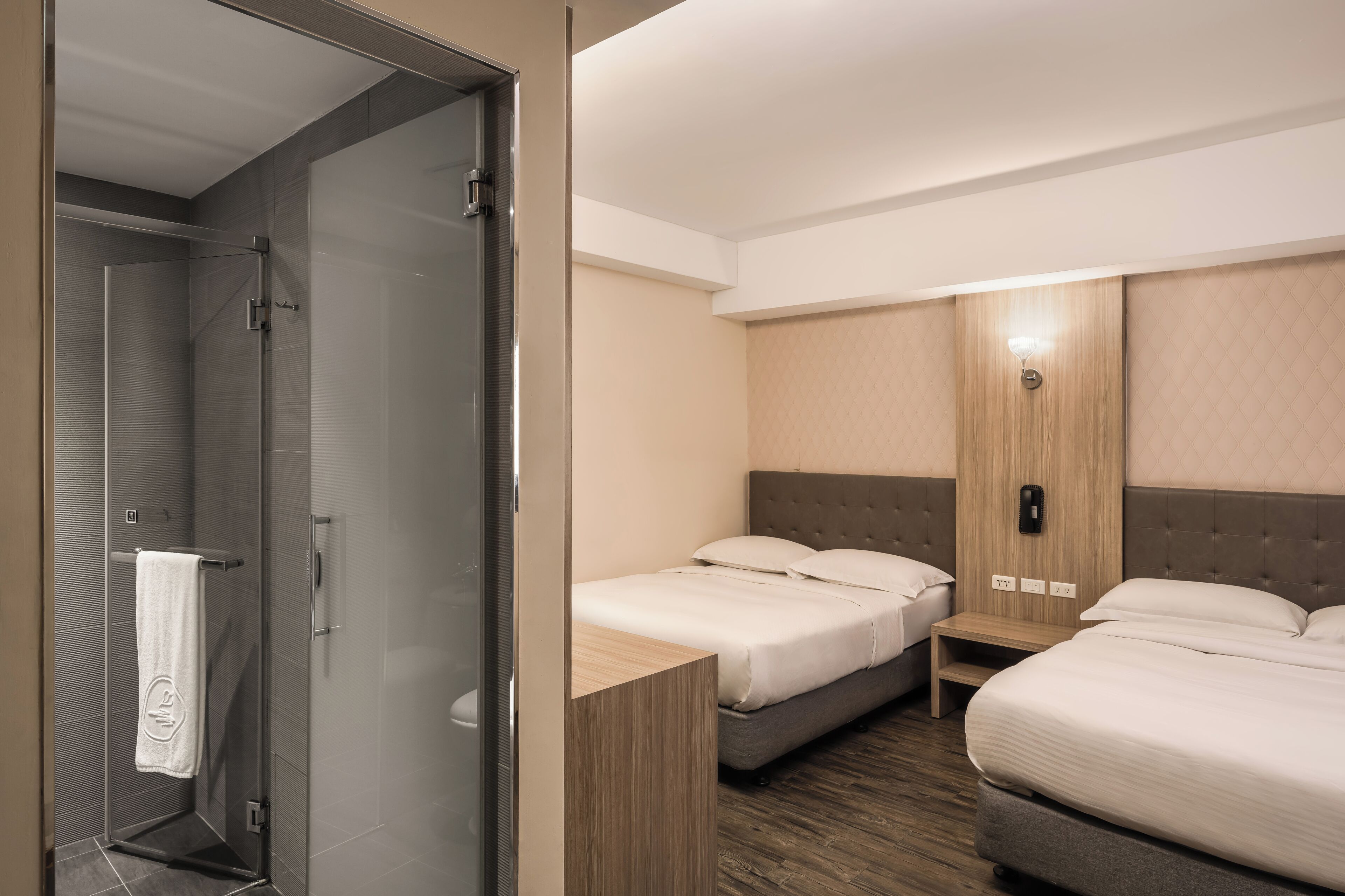 A hotel room with a glass shower enclosure, two beds, a wooden nightstand, and a wall-mounted phone.