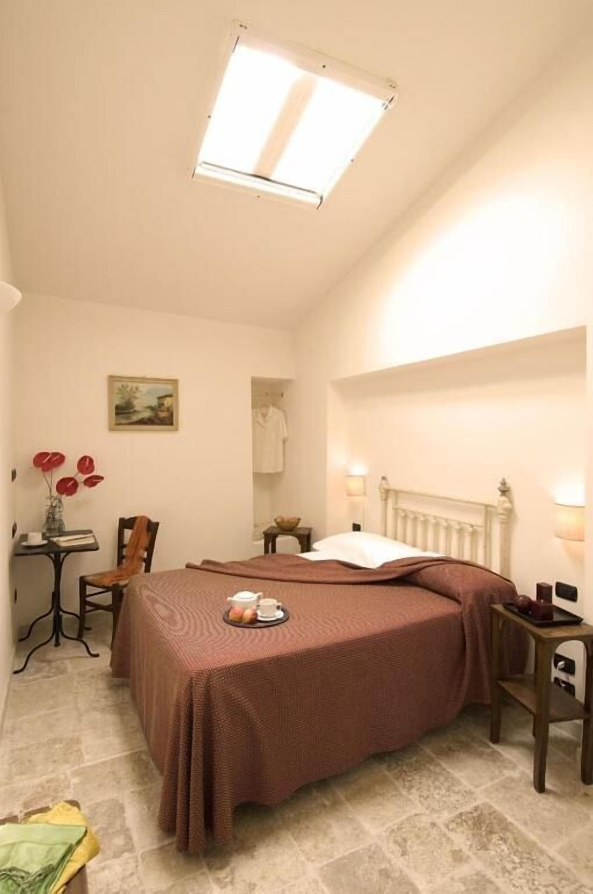 Un dormitorio con una cama, una mesa con un florero, una silla, una mesita de noche con una lámpara y un cuadro en la pared.