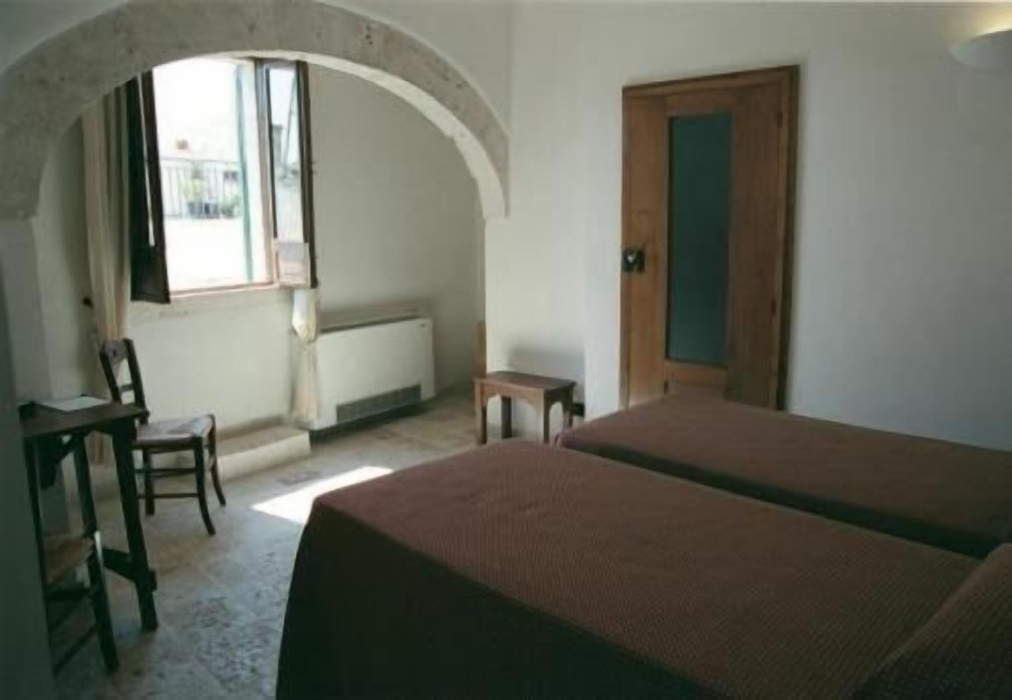 A room with two beds, a wooden door, a small table, a chair, and a window with an arched ceiling.