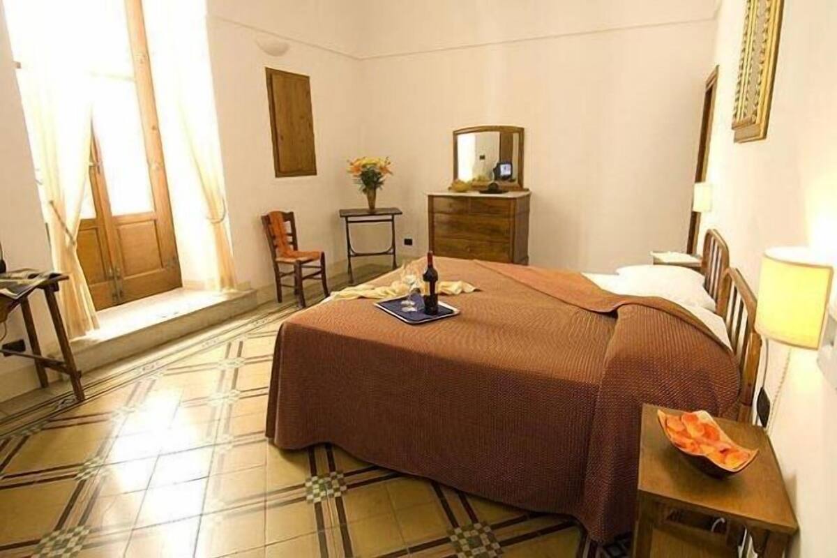 Una habitación de hotel con una cama, un aparador, una silla, una mesita con un jarrón de flores y una ventana con cortinas.