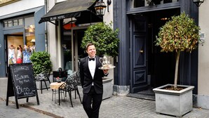 A man in a black suit holding a tray with a teapot and cup, standing in front of a Bliss Hotel with a signboard and outdoor seating area. - Bliss Boutique Hotel (Breda)