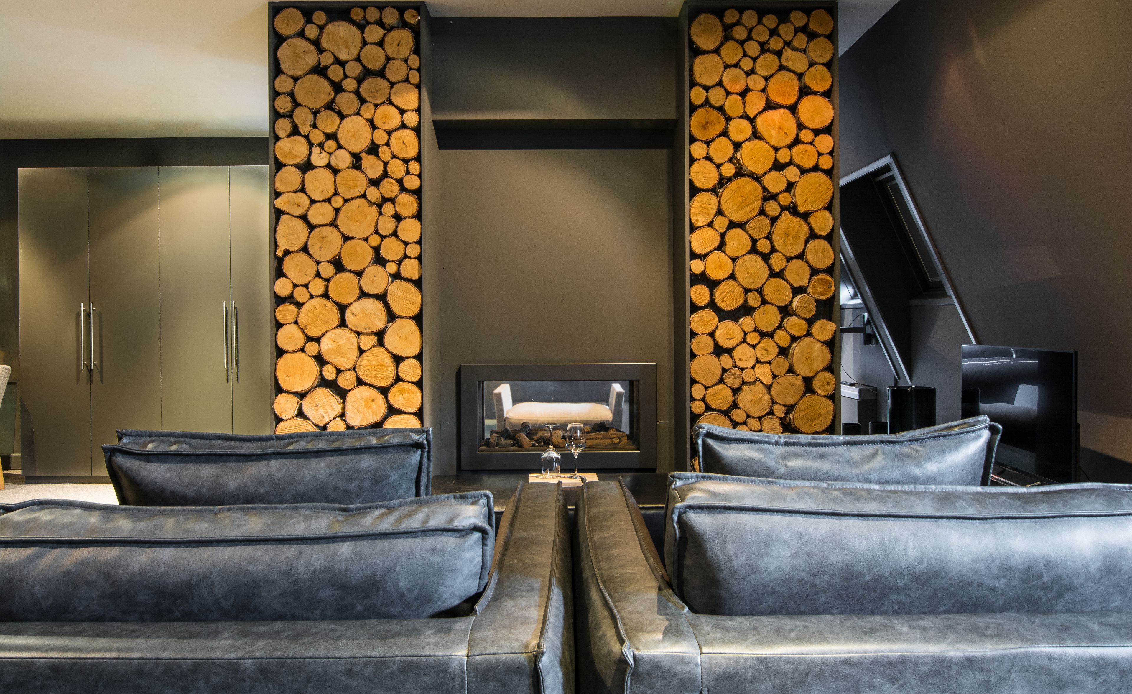 A modern living room with a fireplace, leather sofas, and a wall decorated with stacked logs.