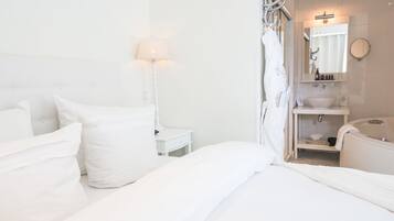 A white hotel room with a bed, bedside table, lamp, and a bathroom with a sink, mirror, and towel rack.