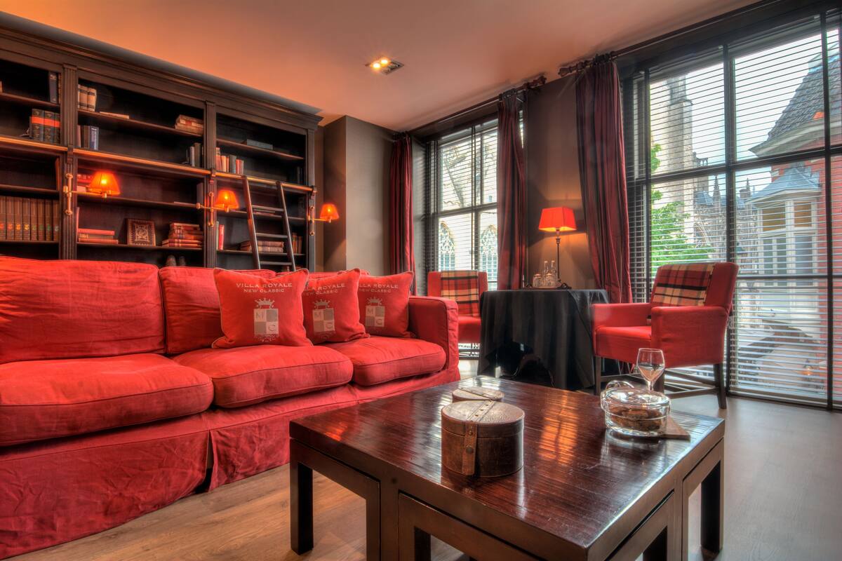 Ein rotes Sofa mit passenden Kissen, ein Holztisch mit einer Glasvase, ein Bücherregal mit Büchern und Dekorationsobjekten sowie ein Fenster mit Jalousien.
