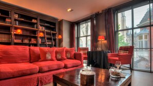 A red sofa with matching cushions, a wooden coffee table with a glass container, a bookshelf with books and decorative items, and a window with blinds. - Bliss Boutique Hotel (Breda)