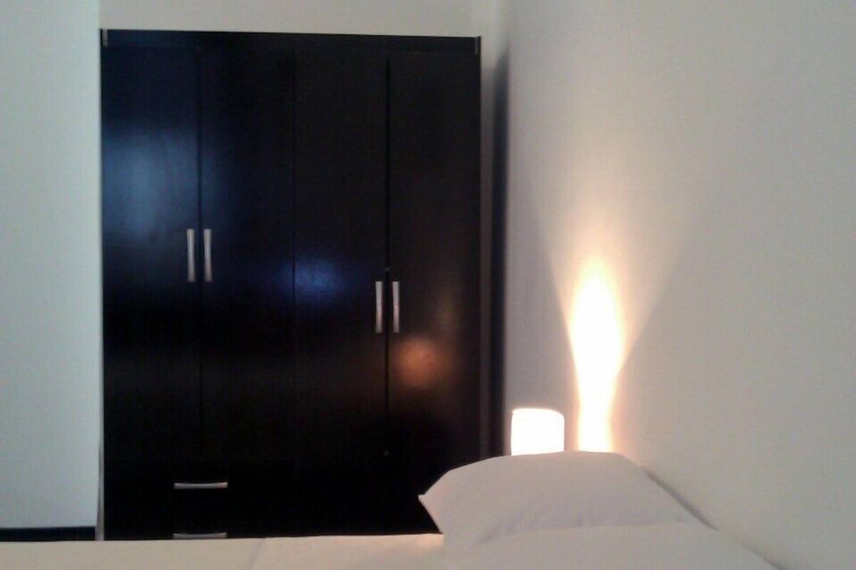 Quarto com cama, guarda-roupa escuro e luminária fixada na parede.