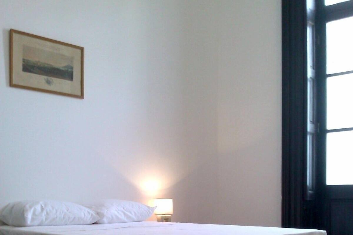 Cama de solteiro num quarto com chão de madeira, luminária fixada na parede e quadro emoldurado.