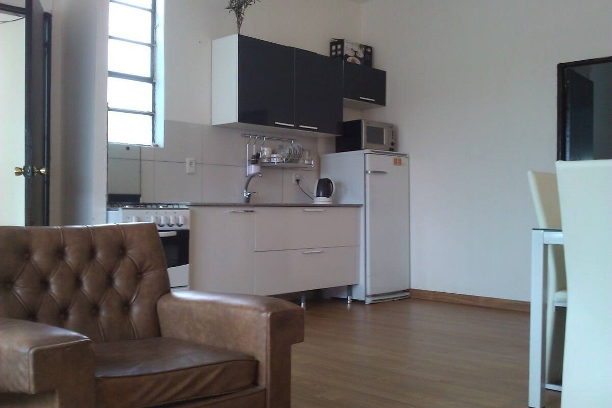 Cozinha moderna com armários brancos, micro-ondas e geladeira. Na sala de estar, um sofá de couro marrom e uma cadeira branca.