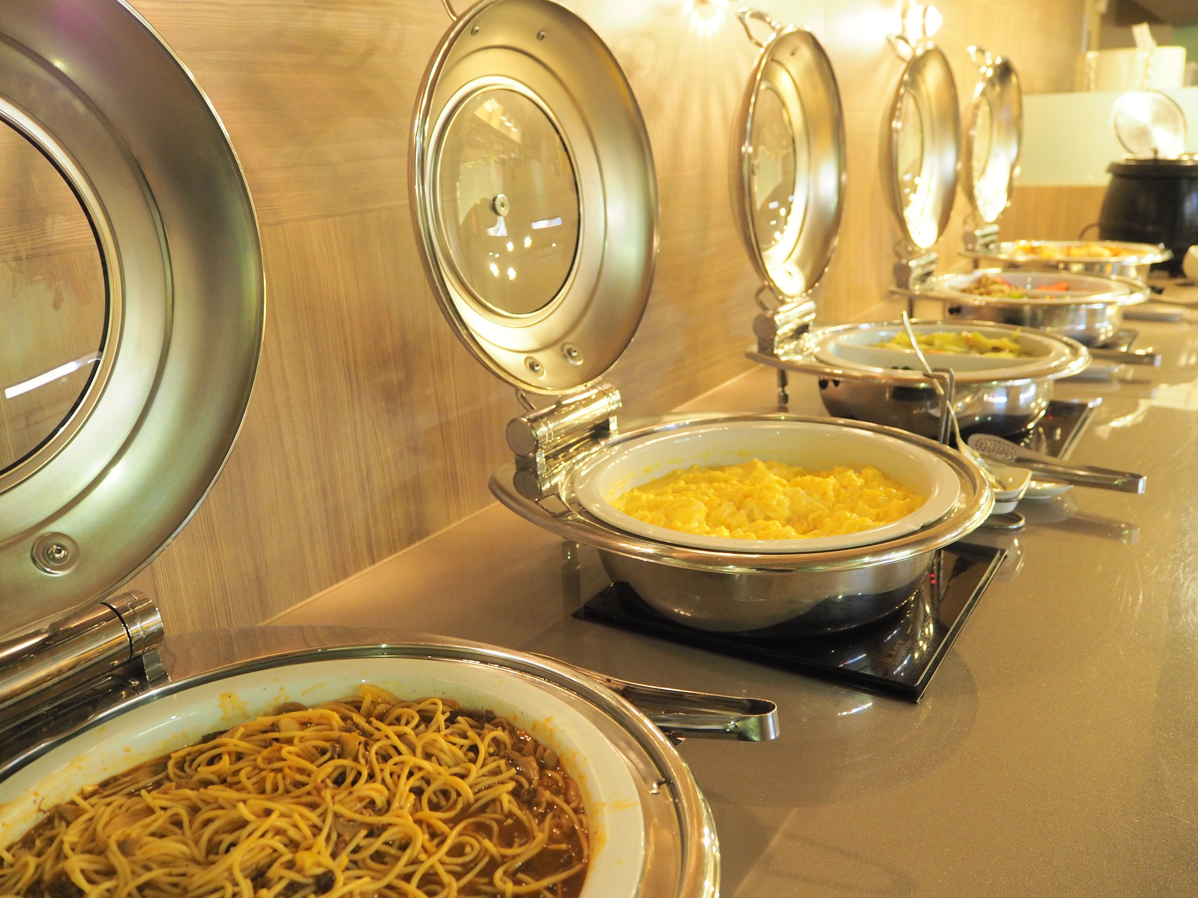 A buffet line with various dishes in metal serving dishes, including noodles and scrambled eggs.