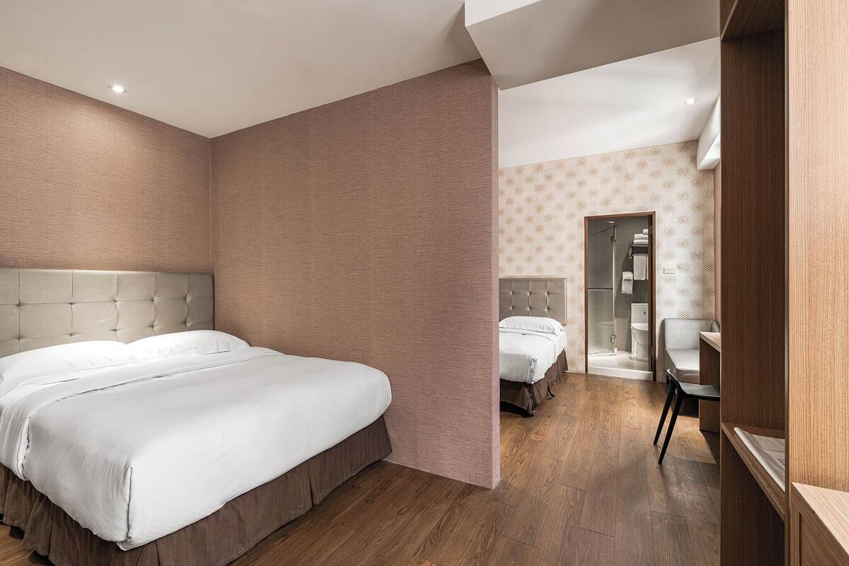 A modern hotel room with a large bed, a smaller bed, a desk, and a shower area.