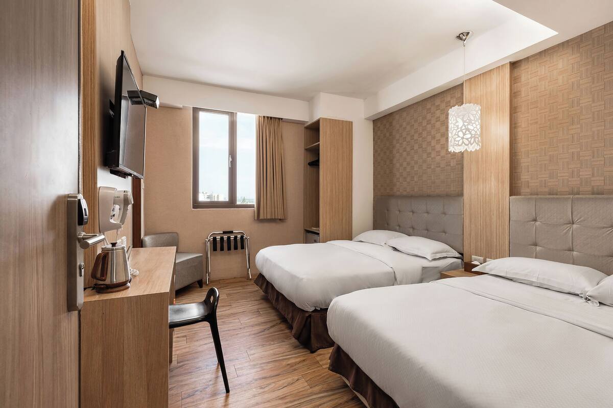 A hotel room with two beds, a desk with a kettle, a chair, a TV, and a window with curtains.