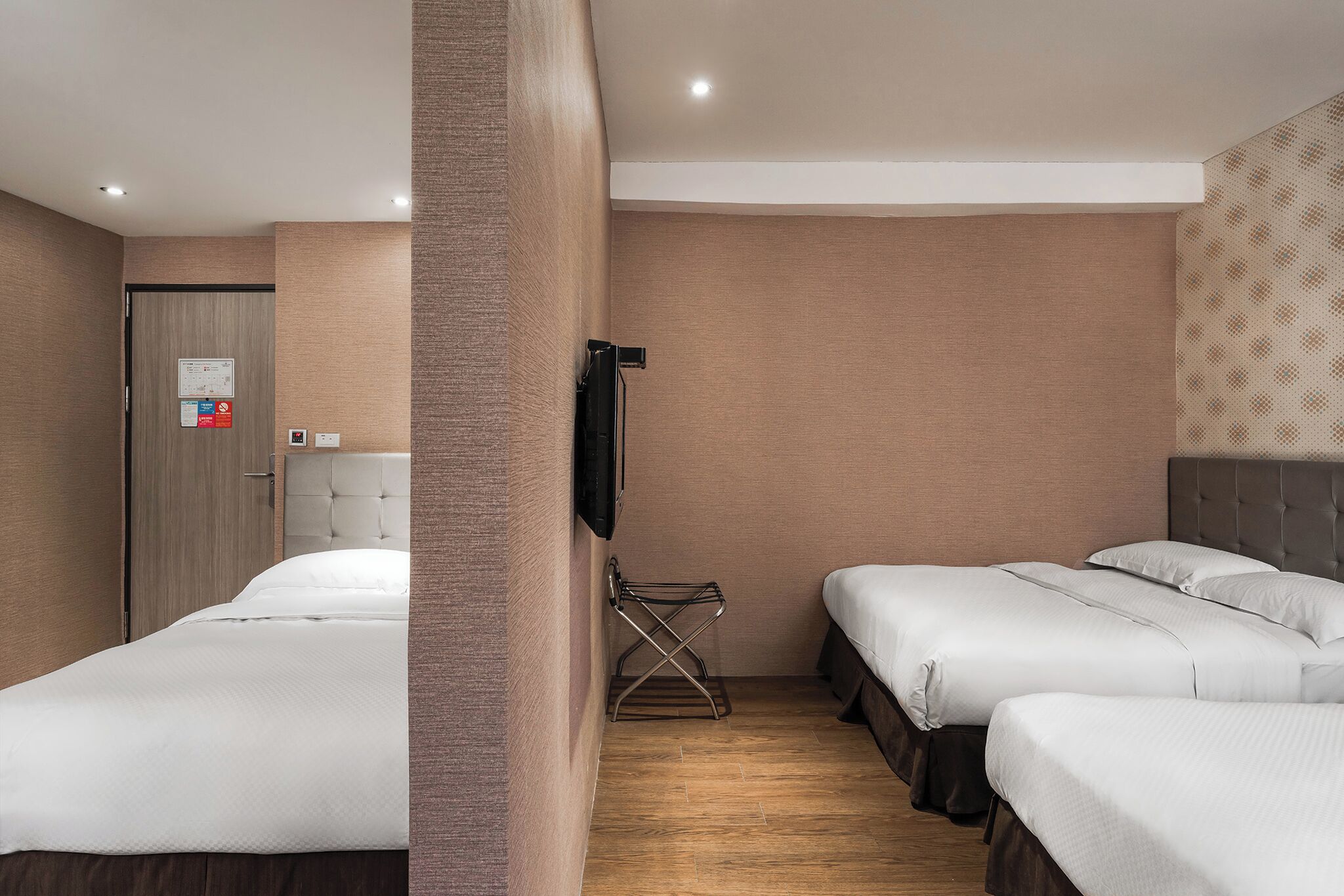 A hotel room with two beds, a wooden floor, a wall-mounted TV, and a door with a sign.