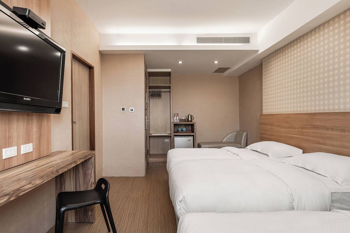 A hotel room with a TV mounted on the wall, a desk with a chair, two beds, a closet, and a small kitchenette.