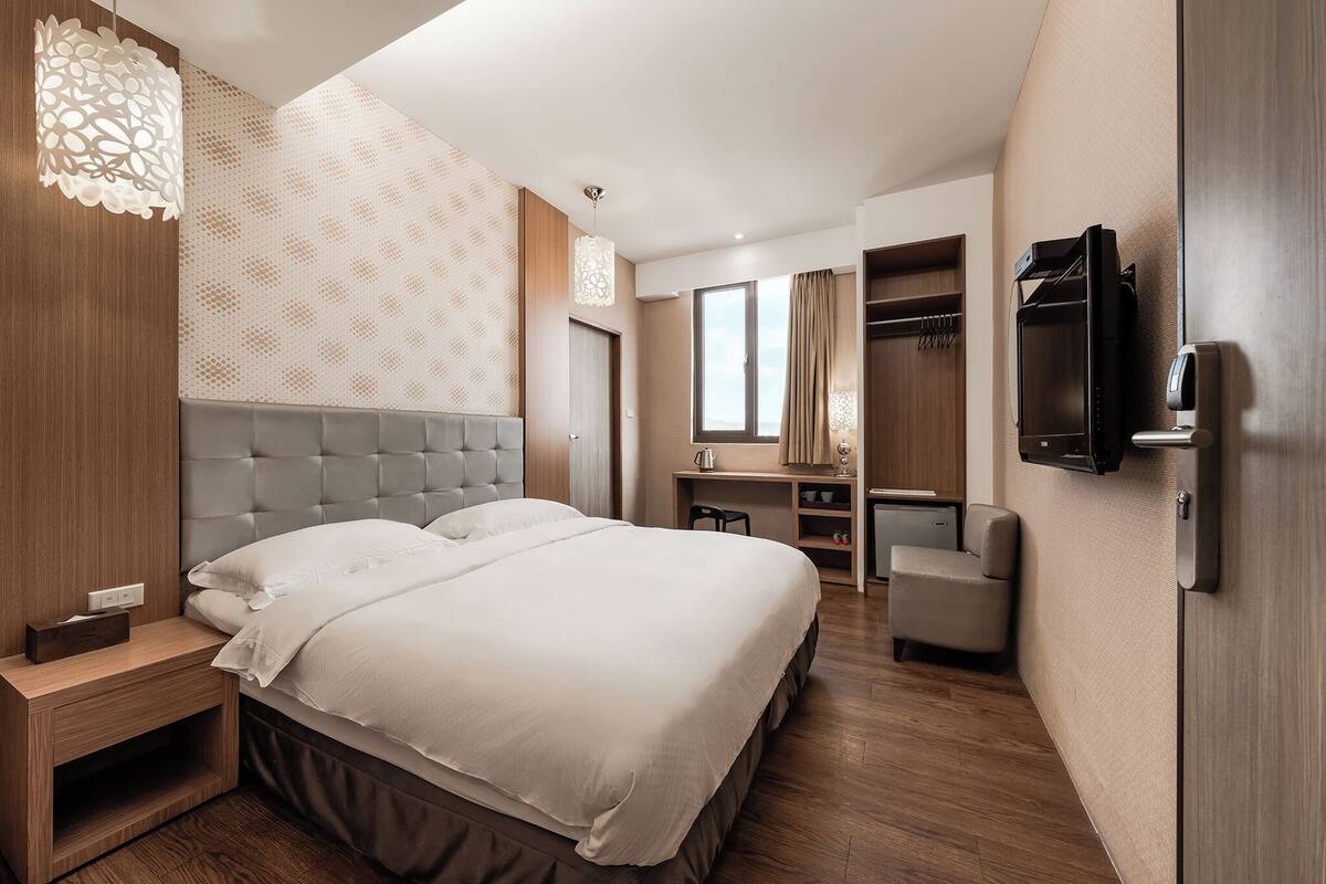 A modern hotel room with a large bed, a desk, a chair, a television, and a wardrobe.