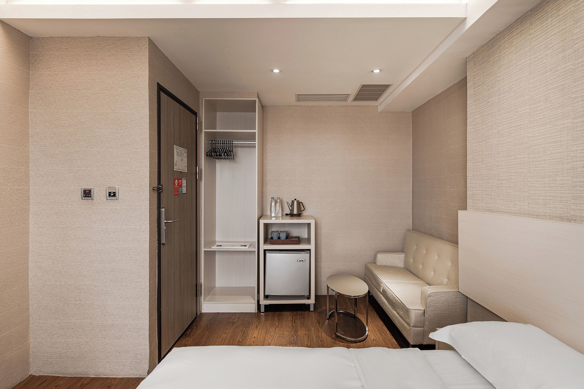 A hotel room with a bed, a seating area with a sofa, a small table, a wardrobe, and a mini-fridge.