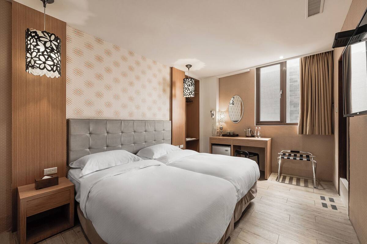 A hotel room with two beds, a desk, a mirror, and a small stool.