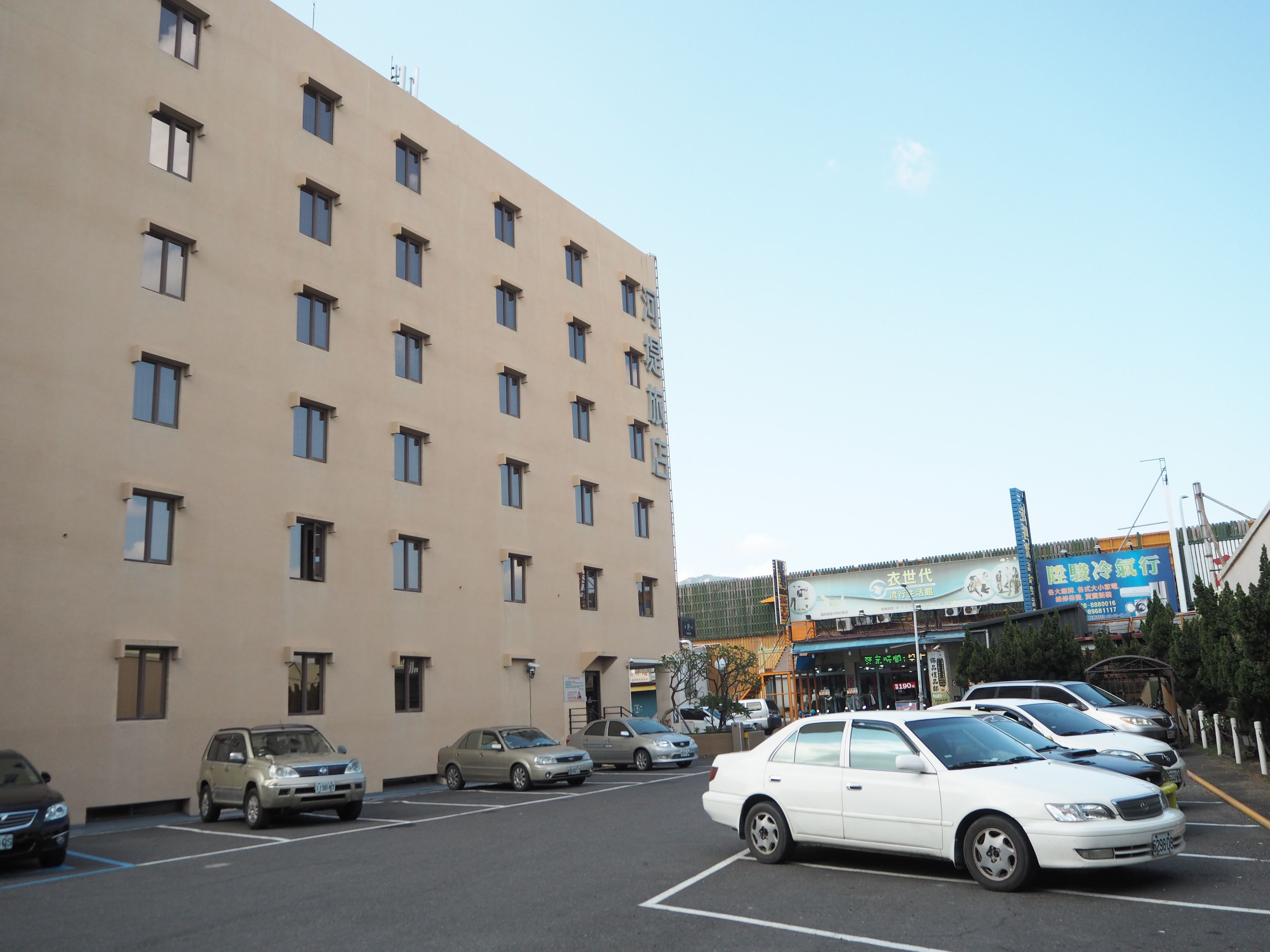 Un edificio de varios pisos con numerosas ventanas, un estacionamiento con varios coches y una zona comercial con señalización en un idioma extranjero.