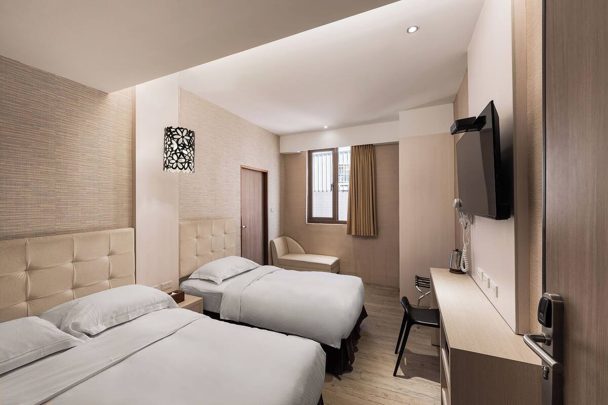 A hotel room with two beds, a desk, a television, and a window with curtains.