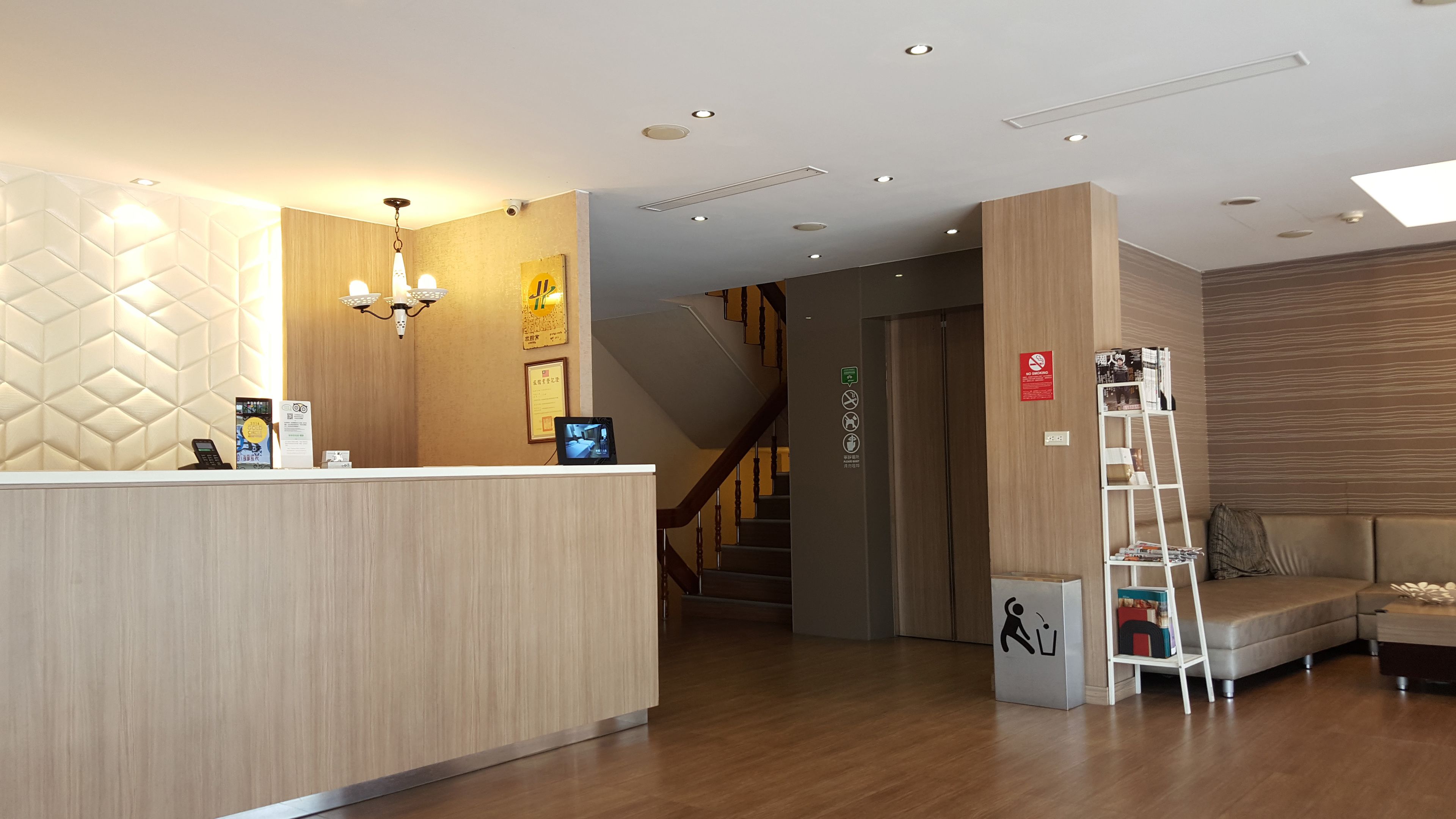A hotel reception area with a wooden reception desk, a chandelier, a digital screen, and a staircase leading to another floor.