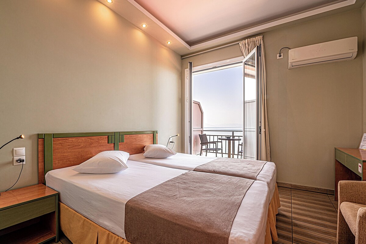 A hotel room with two beds, a nightstand, a desk, and a balcony with a table and chairs.