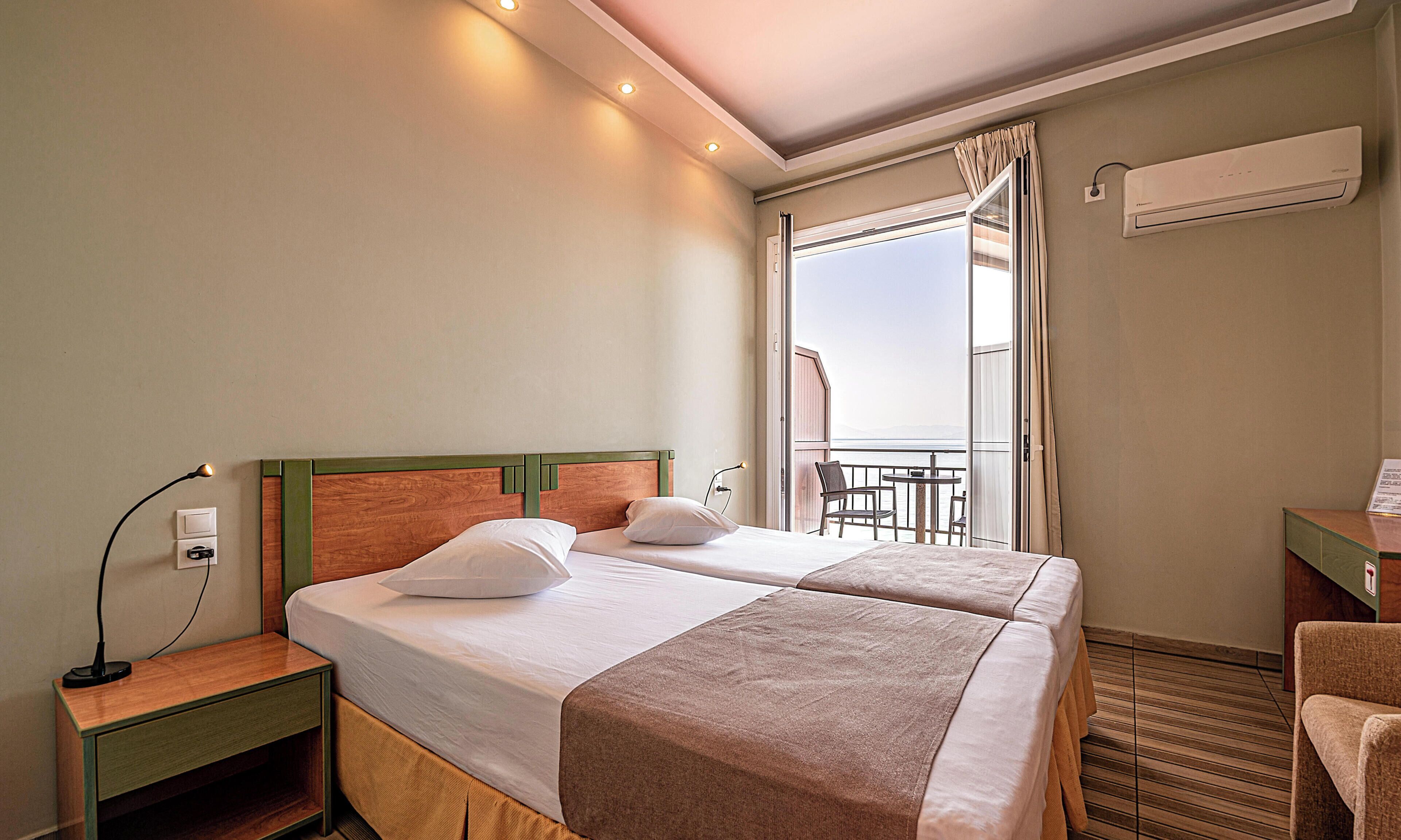 Ein Hotelzimmer mit zwei Betten, einem Nachttisch, einer Lampe, einer Klimaanlage und einem Balkon mit Tisch und Stühlen.