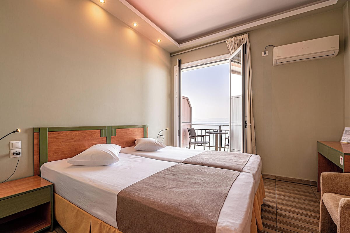 Habitación de hotel con dos camas, una mesita de noche, una lámpara, aire acondicionado y un balcón con mesa y sillas.