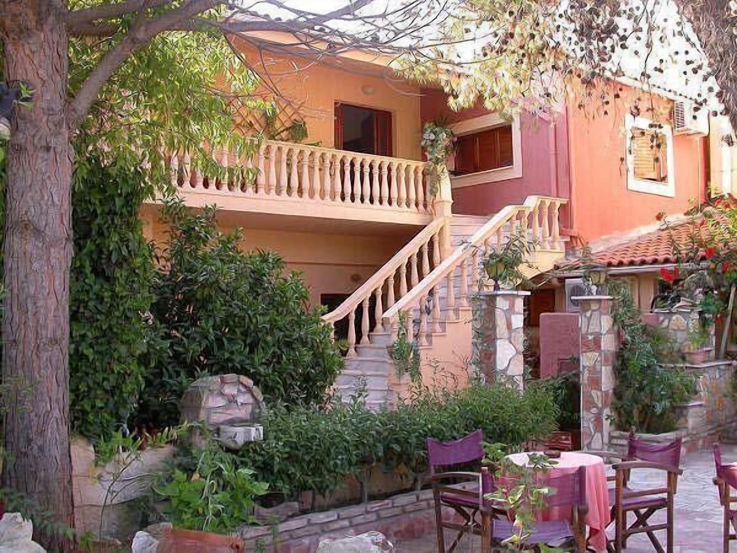 A two-story building with a balcony, a staircase, and a garden area with plants and a table with chairs.