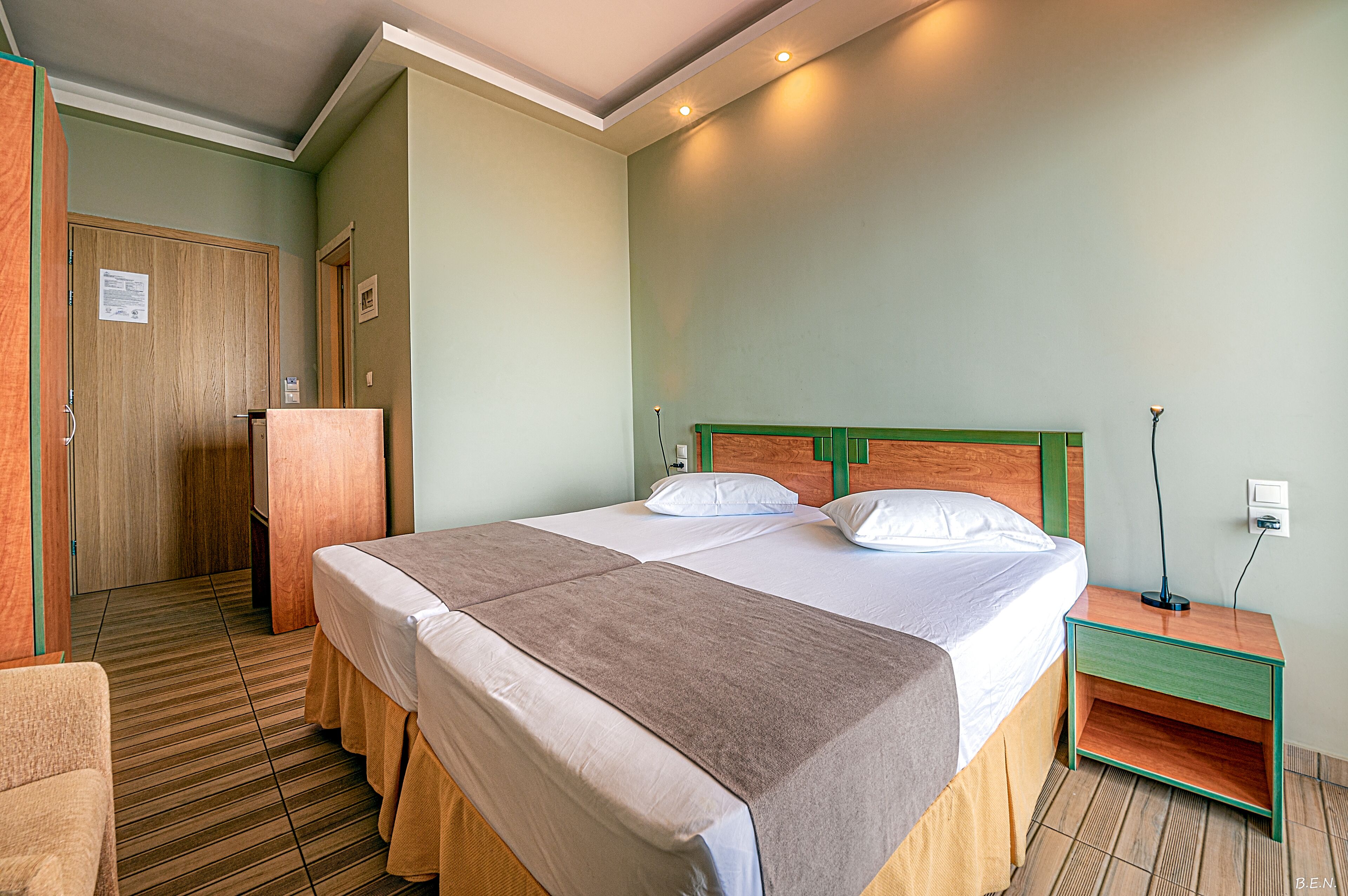 A hotel room with a double bed, a wooden wardrobe, a small bedside table, and a chair.