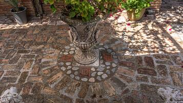 A stone-paved courtyard with a decorative planter in the center, surrounded by brick walls and potted plants.