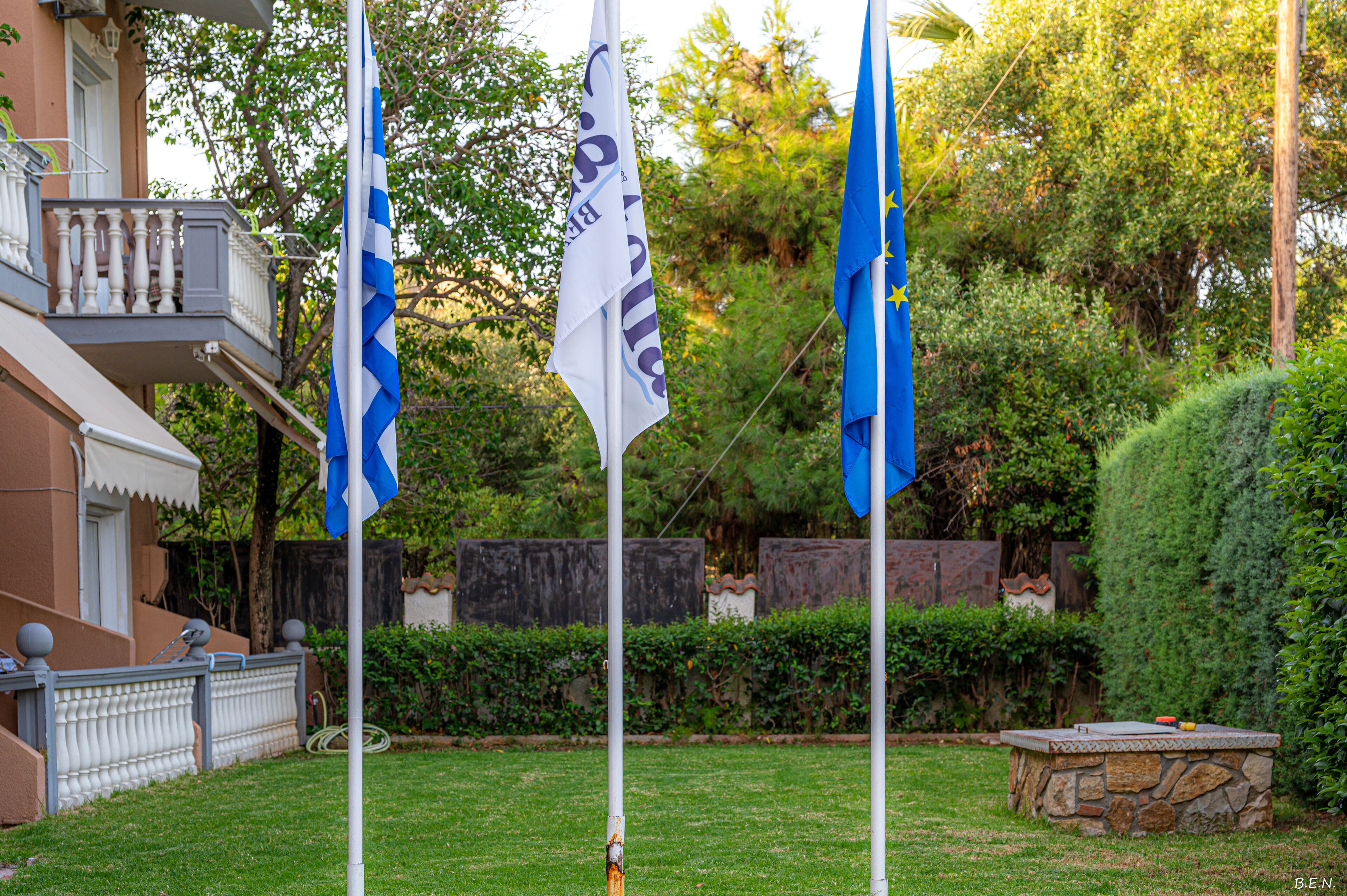 A flagpole with flags, a stone structure, and a building with a balcony.