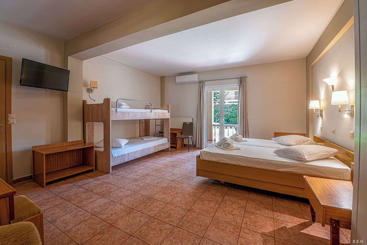 Quarto de hotel com cama de solteiro, uma cama simples, escrivaninha, cadeira e televisão.