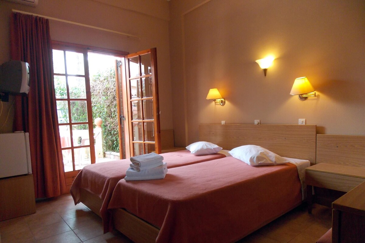 A hotel room with two beds, a wooden headboard, two bedside lamps, a small table, and a window with curtains.