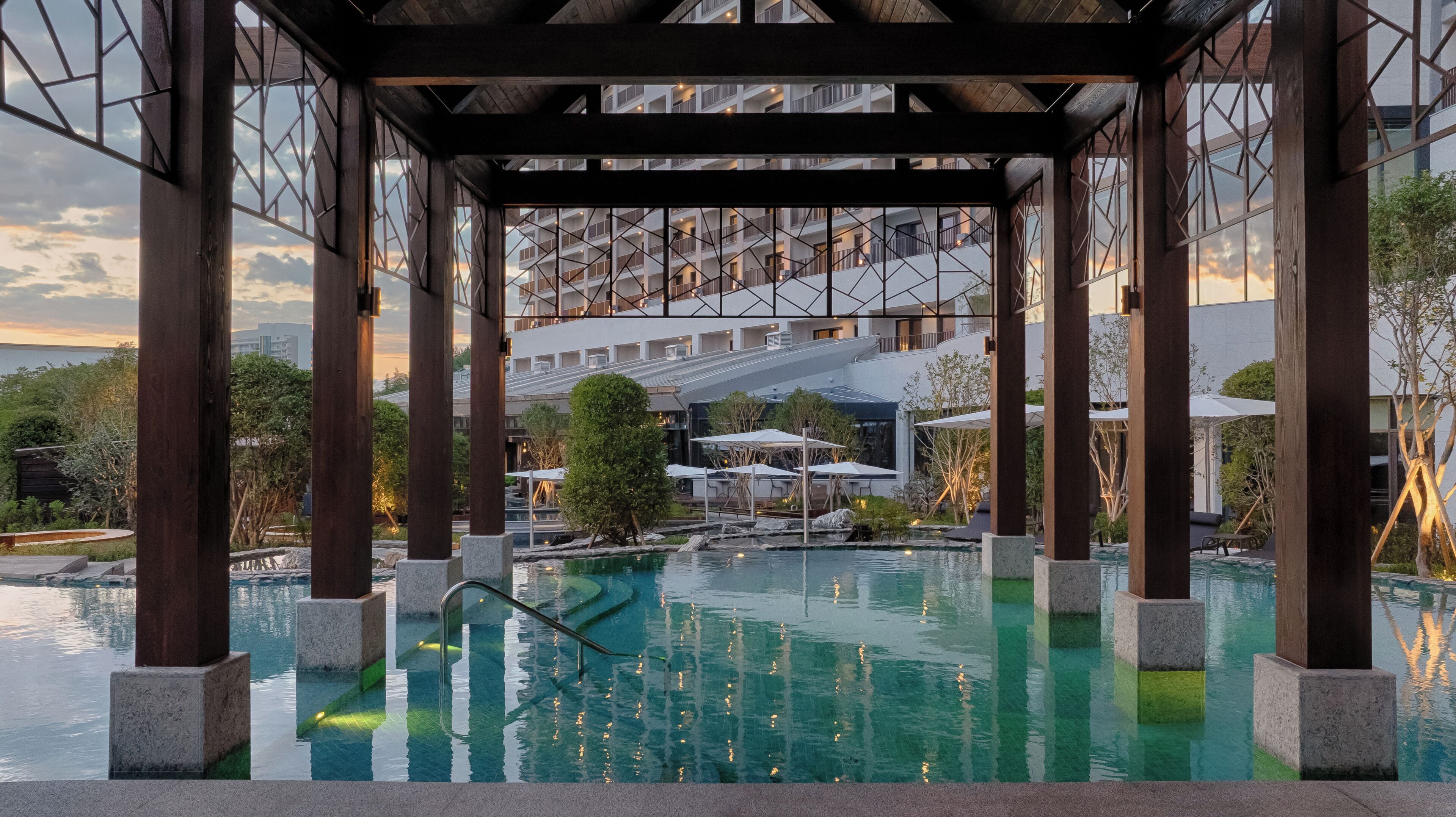 A pool area with a wooden pergola structure, clear pool water, and a view of a modern building with multiple balconies.