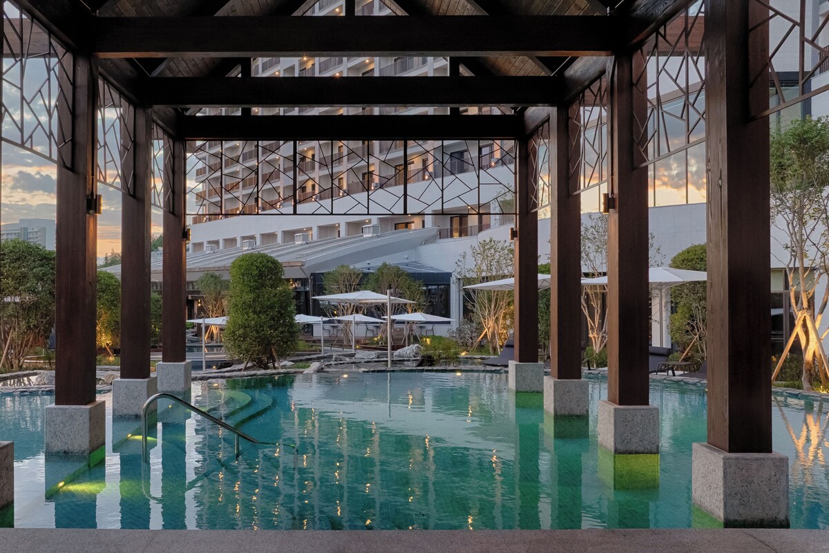 Zona de piscina con una estructura de pérgola de madera, agua cristalina y vistas a un edificio moderno con múltiples balcones.