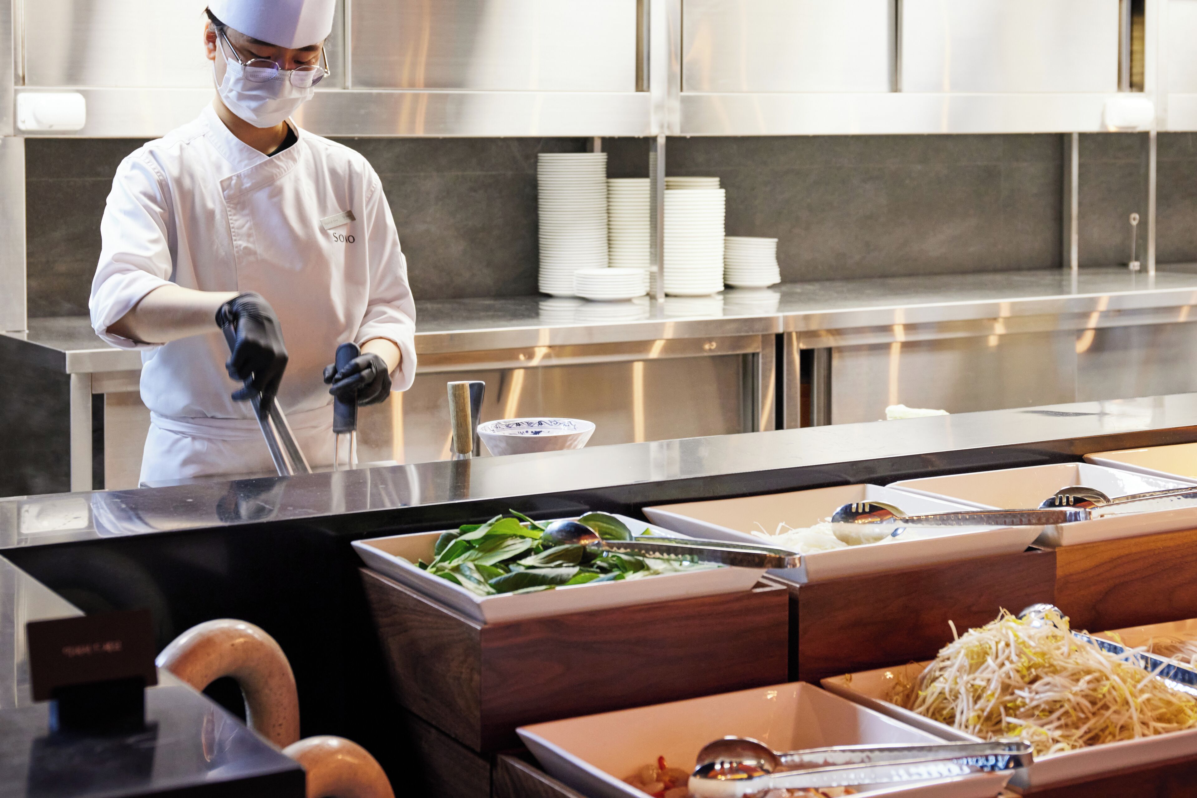 A chef in a white uniform preparing food in a modern kitchen with stainless steel appliances and neatly arranged food stations.