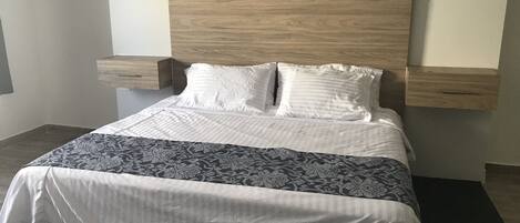 Una cama bien tendida con un cabecero de madera, sábanas blancas y una manta estampada. Hay dos mesitas de noche con lámparas.