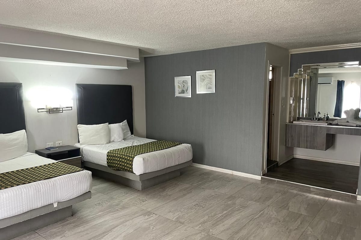Une chambre d’hôtel avec deux lits, une salle de bain avec un miroir et un mur gris.