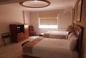 Une chambre d’hôtel avec deux lits, une armoire en bois, une chaise, une petite table et une fenêtre avec des stores.
